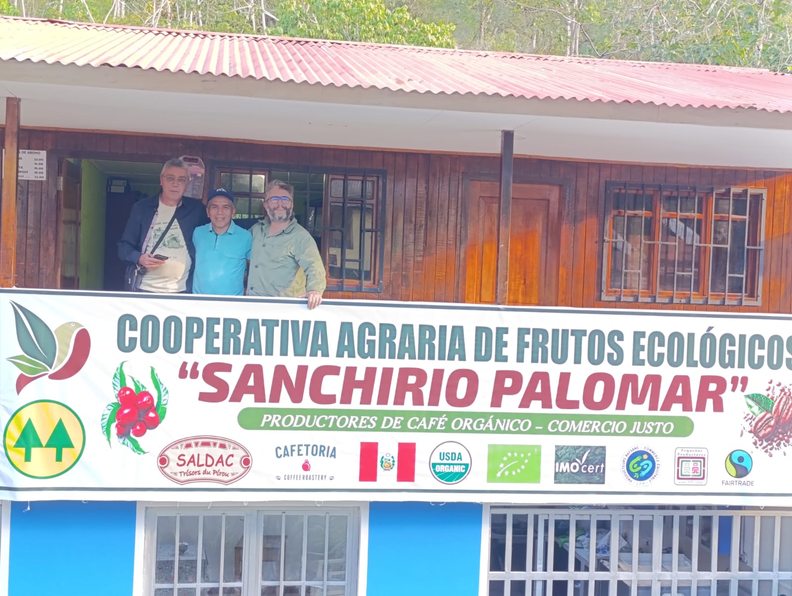 Présidents souriants posant sur le balcon de la Cooperativa Agraria de Frutos Ecológicos "Sanchirio Palomar" au Pérou, devant une grande banderole affichant les certifications bio et commerce équitable
